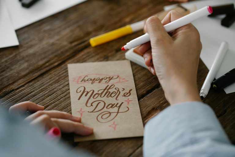 Close-up of a person designing a Mother's Day card with calligraphy on wooden table.